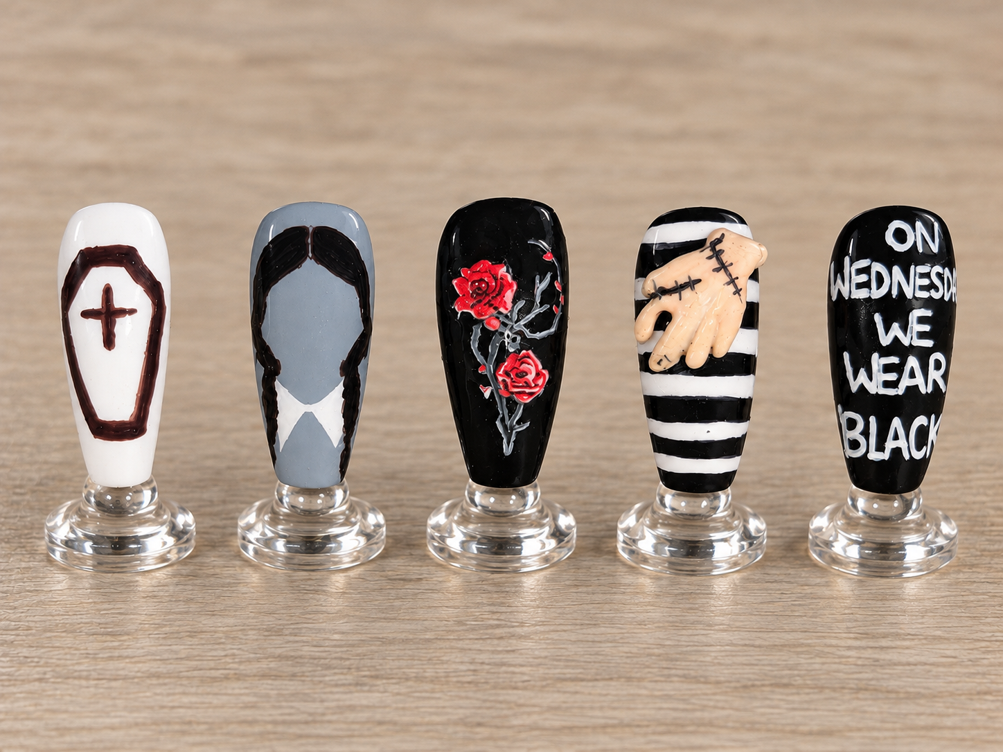 Five Addams Family inspired nails with a coffin, Wednesday silhouette, red roses, a raised Thing hand, and hand painted lettering displayed on clear stands