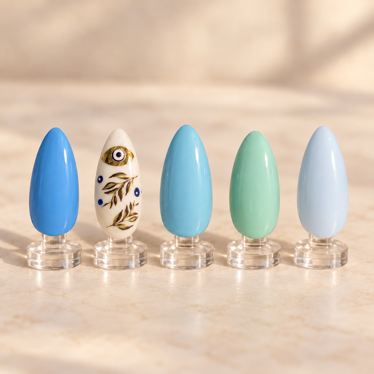 Blue and turquoise nail art with a gold botanical evil eye accent displayed on clear stands
